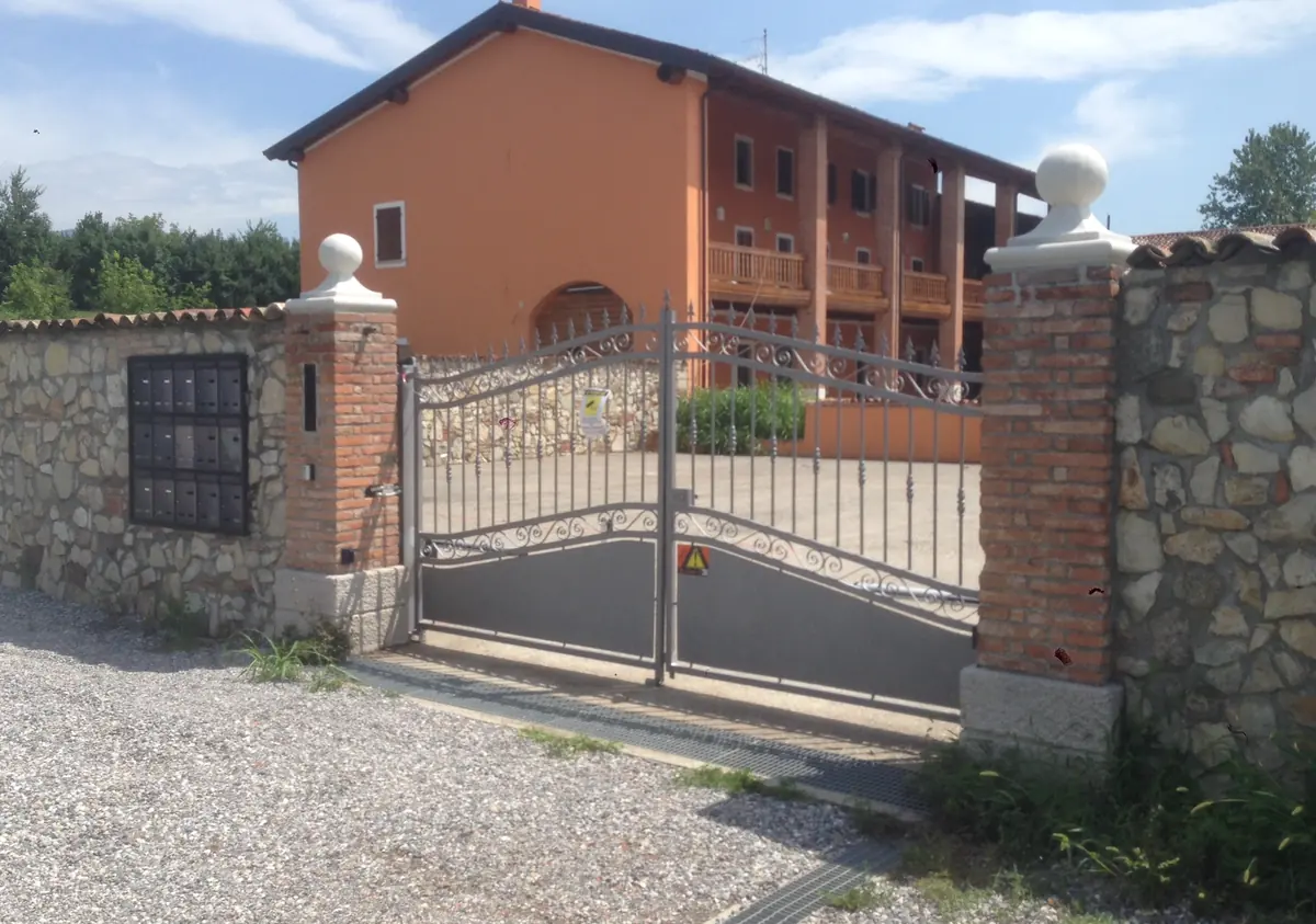 Cancello automatico a due ante battenti in ferro, tra pilastri in mattoni, davanti a una casa di campagna